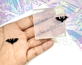 Puede incluir: Un molde de silicona transparente con múltiples formas de murciélagos. El molde está en una mano, y un murciélago negro está al lado.