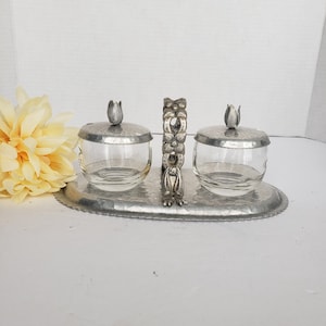 May include: A condiment server set featuring two clear glass bowls with hammered metal lids, each topped with a small metal tulip finial. The bowls sit on an oval hammered metal tray with a central vertical handle decorated with floral motifs.