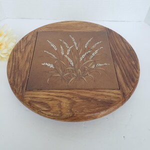 May include: A round wooden serving board featuring a square, brown, matte-finish tile inlay in the center. The tile is decorated with a detailed, light-colored illustration of a cluster of wheat stalks. The wood has a visible grain pattern.
