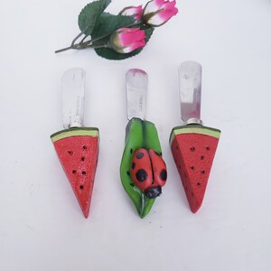 May include: Three watermelon-shaped butter knives with stainless steel blades. The knives have a red and green watermelon design with black seeds. One knife has a ladybug on a green leaf.