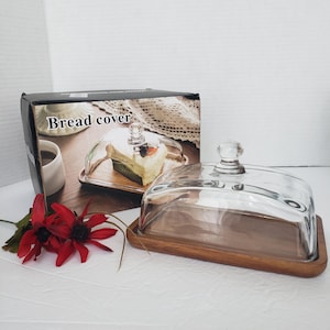 May include: A rectangular bread cover featuring a clear glass dome with a small knob handle, resting on a natural brown wooden base. The product is displayed next to its original cardboard packaging, which shows the text Bread cover.