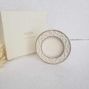 May include: A round, cream-colored ceramic frame with a raised scrollwork border and a thin gold rim. The frame is positioned in front of a square, off-white Lenox American by Design box featuring the brand name and logo printed in black text.
