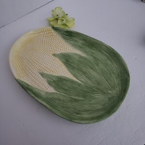 May include: An oval ceramic serving platter shaped like a corn cob. The center features a textured, pale yellow surface resembling corn kernels, while the surrounding area is molded and painted with green, leaf-like patterns with fine lines.