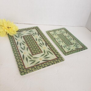 May include: Two rectangular glass cutting boards featuring a light green background with a dark green leaf and yellow flower pattern. The boards have a dark green border with a small floral print and a central rectangular panel of green foliage.