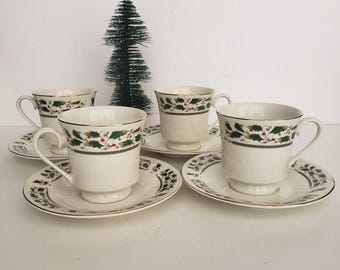 May include: Four white teacups and saucers with a green holly and red berry design. The rims are trimmed with gold. A small green Christmas tree is in the background. The teacups are on a white surface.