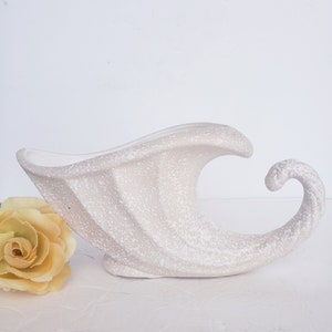 May include: A white ceramic cornucopia vase with a textured surface. The vase has a curved spout and a handle.
