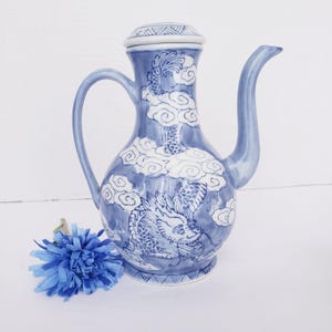 May include: A blue and white ceramic pitcher with a lid, featuring a dragon and cloud design. The pitcher has a curved handle and a long spout. A blue flower is in the foreground. The pitcher is likely for decorative use.