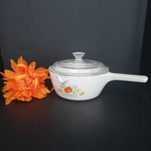 May include: A white ceramic saucepan with a long handle and a clear glass lid. The exterior features a floral pattern with orange, yellow, and green colors. A bright orange artificial flower is positioned next to the cookware item.