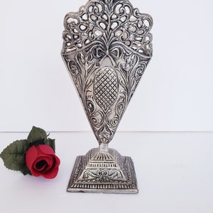 May include: Silver metal vase with an ornate design. The vase has a square base and a tall, slender body with a decorative top. The vase is made of metal and has a silver finish.