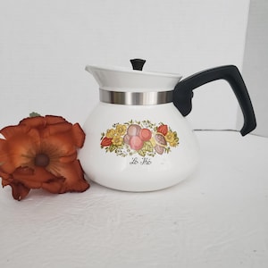 May include: A white teapot with a black handle and lid, featuring a metallic band around the neck and a colorful botanical illustration of fruits and flowers on the front. The text "Le Thé" is printed in black script below the fruit design.