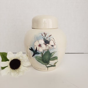 May include: White ceramic jar with a lid, featuring a floral design with white flowers and a hummingbird in flight. The jar has a blue and green background with the text "Gardenia East Indies" at the bottom.