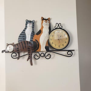 May include: A whimsical cat-themed wall clock. The clock features three stylized cat figures in brown, gray, and orange, with a round clock face. The clock is mounted on a decorative black metal bracket.