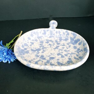 May include: An oval ceramic dish featuring a white base with a speckled light blue pattern and a small handle at the top. A single artificial blue flower with a green stem is positioned to the left of the dish, resting on a dark surface.