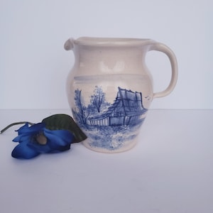 May include: A white ceramic pitcher with a blue and white hand-painted scene of a house and trees. The pitcher has a wide mouth and a handle.