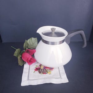 May include: A white teapot with a rounded body, a metallic silver lid, and a dark gray handle. The teapot sits on a white square fabric coaster featuring an embroidered basket of fruit, accompanied by a single artificial red rose with green leaves.