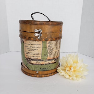 May include: A vintage wooden cylindrical box with a black handle and metal clasp. The box is adorned with a map design and text in Latin. A cream-colored flower rests beside the box, adding a touch of elegance. The box is a unique storage item.