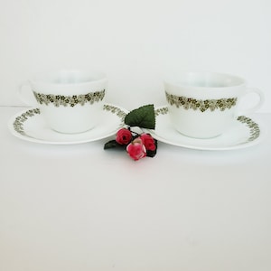 May include: Two white teacups with green floral patterns and matching saucers. The cups and saucers are arranged in a heart shape with a small bouquet of red roses in the center.