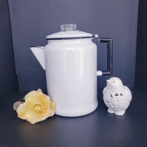 May include: A white enamel coffee percolator with a black handle, a silver-toned rim, and a clear glass knob on the lid. Beside the percolator sits a pale yellow fabric rose and a small white ceramic bird figurine with intricate cut-out details.
