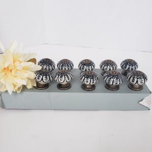 May include: A set of ten round ceramic cabinet knobs featuring a white base with a dark blue leaf-like pattern and a bronze-toned metal base. The knobs are arranged in two rows on a rectangular gray box, accompanied by a large cream-colored flower.