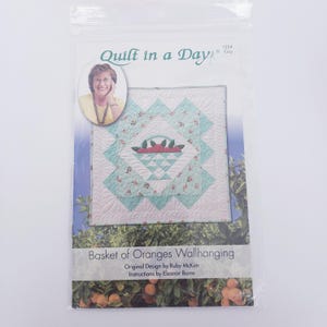 May include: A package for a quilt project titled "Basket of Oranges Wallhanging." The design features a quilted basket filled with red fruit, surrounded by a turquoise and floral patterned border. The package includes instructions by Eleanor Burns.