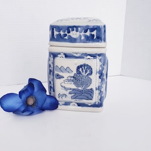 May include: A square, lidded ceramic jar with a blue and white landscape design. The jar features a detailed illustration of a tree, mountains, and houses. The lid and sides have floral and geometric patterns. A blue flower rests near the jar.