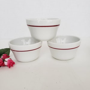 May include: Three white ceramic bowls with a dark red band around the top. The bowls are stacked in a pyramid shape. The bowls are on a white surface.
