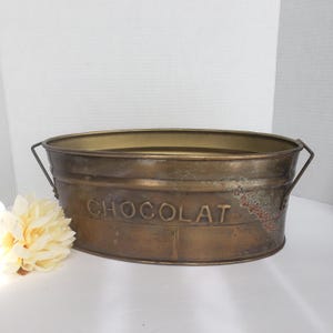 May include: An oval-shaped brass basin with two side handles and the word CHOCOLAT embossed on the front. The metal surface features a weathered, aged patina with visible oxidation and dark spots throughout the exterior of the container.