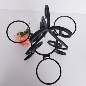 May include: Black metal decorative wine rack with a unique, abstract design featuring multiple circular holders and curved accents. A peach-colored flower adds a touch of color. The rack is designed to hold wine bottles.