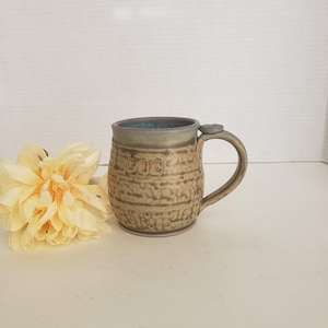 May include: A handmade ceramic mug with a textured, earthy brown exterior and a teal-colored interior. The word "PIC" is embossed on the front. The mug has a rounded handle and a slightly flared rim. A yellow flower is in the background.