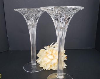 May include: Two clear glass candle holders with a decorative, textured design. The holders have a wide, flared top and a tall, slender stem.