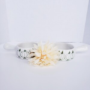 May include: Two white ceramic bowls with handles, each featuring a repeating green line-art pattern of onions around the exterior. A large, cream-colored artificial flower is positioned in the center, partially obscuring the two bowls.