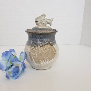 May include: A ceramic jar with a lid featuring a fish-shaped handle. The jar is two-toned, with a blue-gray top and a beige base. The words "spare air" are inscribed on the front.