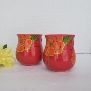 May include: Two red ceramic mugs with orange and yellow floral designs. Each mug has a small handle and a glossy finish. The floral pattern includes green leaves and stems, adding a touch of nature.