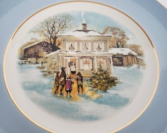 May include: A decorative plate with a light blue border and a gold rim. The center features a winter scene with a house, people, and a Christmas tree. The text "Christmas 1977" is in gold lettering.