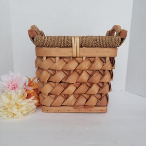 May include: A rectangular woven basket featuring light brown wooden slats arranged in a diagonal pattern. The top rim is wrapped in a thick, textured brown rope, and the basket includes two wooden handles attached to the sides for carrying.