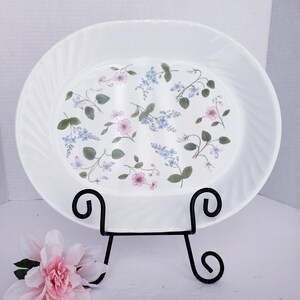 May include: An oval white serving platter with a subtle swirl texture on the rim, decorated with a pattern of small pink and blue flowers and green leaves. The platter is displayed on a black metal stand with curved scrollwork feet.
