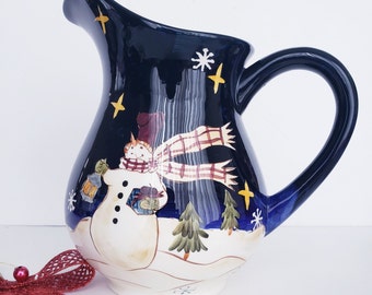 May include: A ceramic pitcher with a dark blue background, featuring a snowman design. The snowman wears a scarf and holds a lantern. The pitcher has a curved handle and a white base, with a red bow in the foreground.