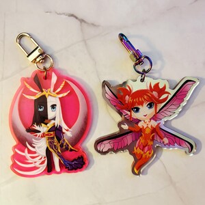 May include: Two acrylic keychains featuring anime-style characters. One keychain has a character with black and white hair, set against a pink crescent moon. The other keychain depicts a fairy with red hair and pink wings. Both have metal clasps.