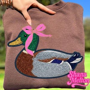 May include: Brown sweatshirt featuring a large duck applique with green, brown, gray, and blue details. The duck has a pink bow around its neck. The sweatshirt has the text "Shana Melton" in pink script.