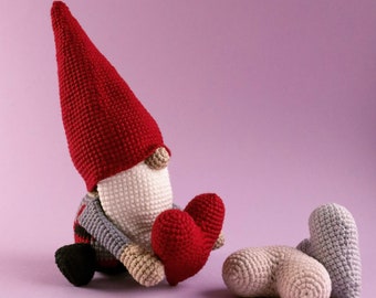 May include: A handmade crochet gnome with a red hat, gray sweater, and holding a red heart. Two additional crochet hearts, one beige and one gray, are next to the gnome. The background is a solid light purple.