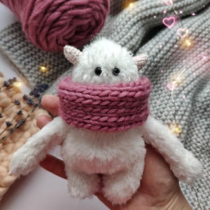 May include: A white fluffy toy with a pink knitted scarf. The toy has large black eyes and small horns. The toy is sitting on a hand.