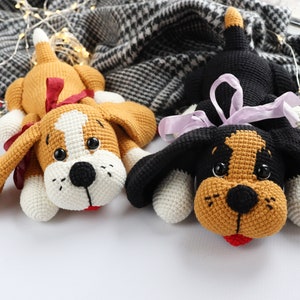 May include: Two crocheted dog plushies, one brown and white with a red bow, and one black and brown with a purple bow. Both plushies have white paws and black noses.