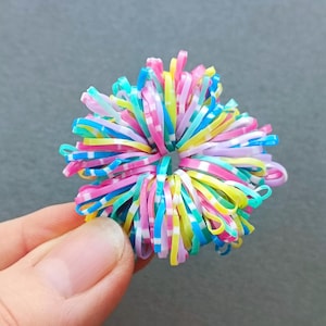 May include: A colorful hair scrunchie made of small, elastic bands in shades of pink, blue, yellow, and white. The scrunchie is round and has a flower-like appearance. The background is a solid gray.