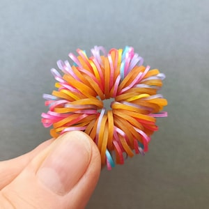 May include: A colorful rubber band ball held by a hand. The ball is composed of numerous rubber bands in shades of orange, pink, yellow, purple, and blue. The bands are arranged in a radial pattern, creating a textured, spherical shape.