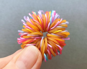 Puede incluir: Una colorida bola de goma sostenida por una mano. La bola está compuesta por numerosas bandas elásticas en tonos naranja, rosa, amarillo, morado y azul. Las bandas están dispuestas radialmente, creando una forma esférica texturizada.