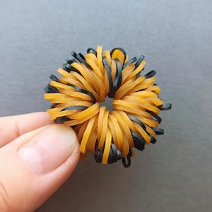 May include: A handmade hair scrunchie made of orange and black rubber bands. The scrunchie is round with a hole in the center. The rubber bands are arranged in a radial pattern, with alternating colors of orange and black.