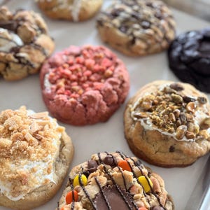 Può includere: Una varietà di biscotti gourmet su una teglia. I biscotti presentano diversi condimenti, tra cui noci, gocce di cioccolato e caramelle colorate. Alcuni biscotti hanno una tonalità rossa, mentre altri sono ricoperti di glassa bianca e briciole.