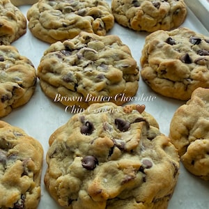 May include: A tray of golden-brown chocolate chip cookies with visible dark chocolate morsels and a light dusting of salt. The text "Brown Butter Chocolate Chip Cookies" is centred on the image, accompanied by a cookie and a heart emoji.