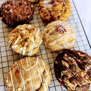 May include: An assortment of gourmet cookies on a wire rack. The cookies feature various toppings, including chocolate drizzle, nuts, and sprinkles. Some cookies have a light-colored base, while others are dark chocolate. The cookies are arranged on a black wire rack.