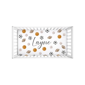 Puede incluir: Una cuna blanca con una sábana bajera con un estampado de balones deportivos, incluyendo balones de baloncesto, fútbol, béisbol y fútbol americano. El nombre "Layne" está escrito en cursiva en la sábana.